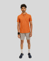Man wearing an orange polo shirt and beige shorts on a gray background