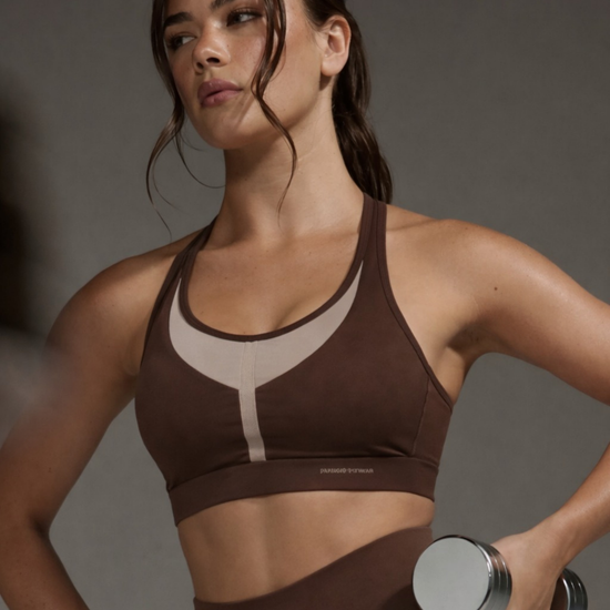 Woman in brown sports bra and leggings holding a dumbbell against a gray background