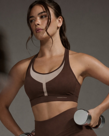Woman in brown sports bra and leggings holding a dumbbell against a gray background
