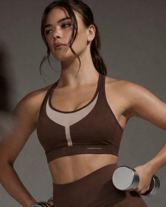 Woman in brown sports bra and leggings holding a dumbbell against a gray background