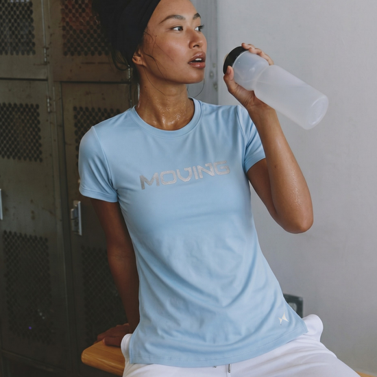 Person wearing a light blue t-shirt with 'MOVING' text, sitting and holding a water bottle.