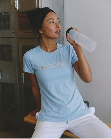 Person wearing a light blue t-shirt with 'MOVING' text, sitting and holding a water bottle.