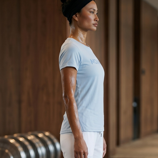Woman in a light blue t-shirt and white pants standing in a yoga pose indoors.
