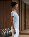 Woman in a light blue t-shirt and white pants standing in a yoga pose indoors.