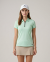 Woman wearing a light green polo shirt and beige skirt with an Adidas cap on a plain background