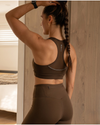 Woman wearing a brown sports bra and leggings, standing in a room with wooden walls.