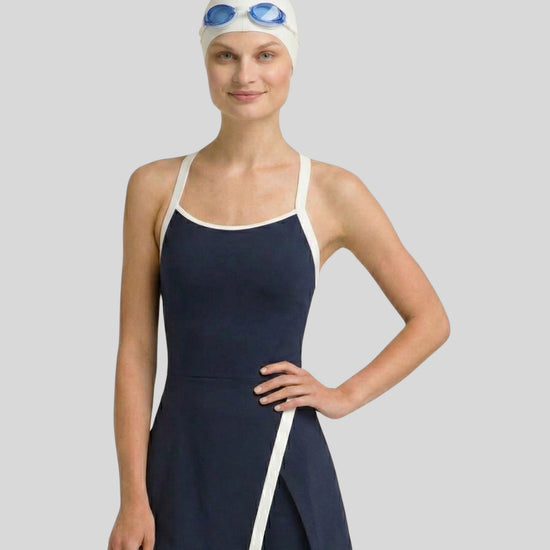 Woman wearing a navy swimsuit with white trim, white swim cap, and blue goggles on a gray background