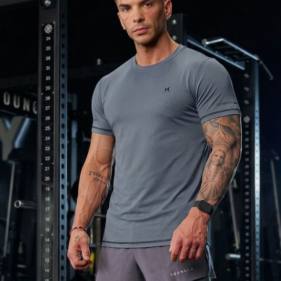 Man wearing a gray athletic outfit with tattoos on his arms and legs, standing in a gym setting.