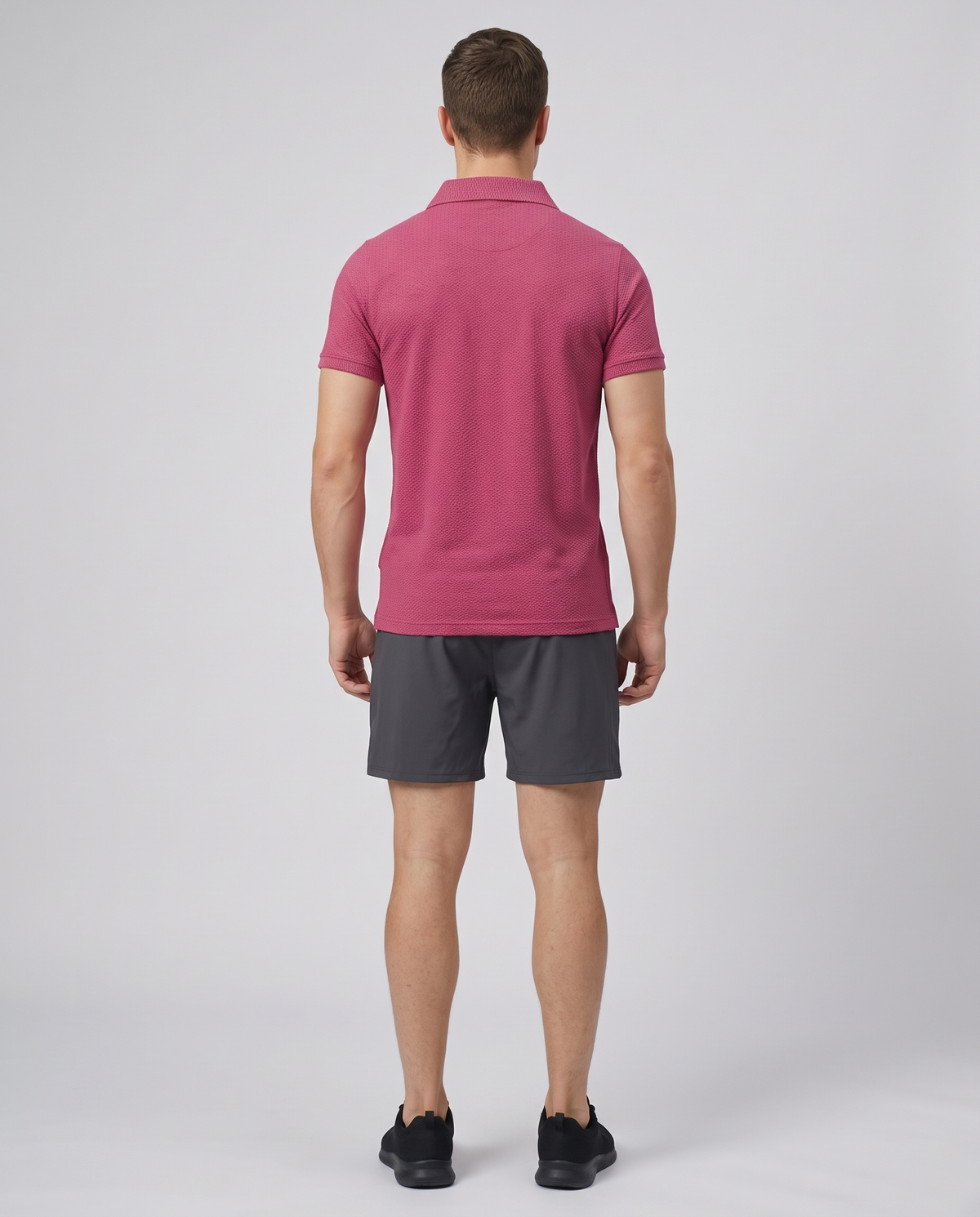 Man wearing a wine textured bubble-knit polo shirt and charcoal shorts, back view on a pale grey studio background
